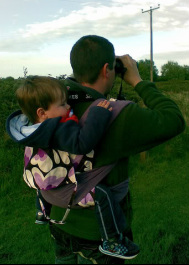 A dad looks through binoculars while carrying his son in a soft structured carrier on his back